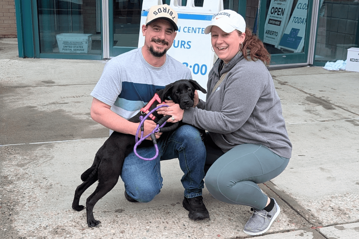 Two people smiling with a black puppy outside the shelter