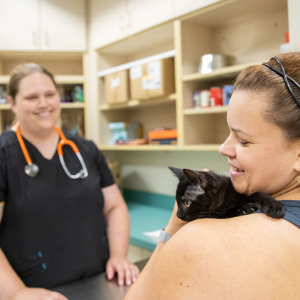 A veterinarian smiles at a client who is holding a black kitten on her shoulder